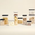 Un groupe de boîtes de conservation transparentes avec des couvercles noirs et blancs, remplies de diverses pâtes, céréales, haricots, noix et cacahuètes.