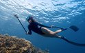 Vrouw in zwart duikpak met flippers filmt onder water bij koraalrif.