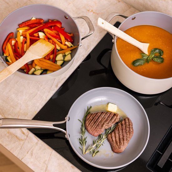 Grijze koekenpan met twee biefstukken en rozemarijn, naast een pan met oranje soep, op een inductiekookplaat.