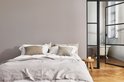 Een opgemaakt bed met beige en taupe kussens en een lichtgrijs dekbed, naast een houten krukje en een glazen deur naar een badkamer.