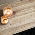 Close-up van het licht houten tafelblad met een grof patroon. Twee glazen windlichten met brandende kaarsen.