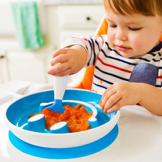 Een baby met gestreepte shirt eet pompoenpuree uit een blauw smiley bord met zuignap, met behulp van een blauw-witte lepel.