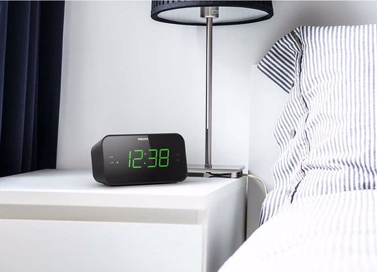 Zwarte Philips wekkerradio met groene digitale cijfers, op een nachtkastje naast een bed.