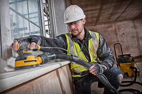 Man in veiligheidsuitrusting schuurt een vensterbank met een gele en zwarte DeWalt schuurmachine.