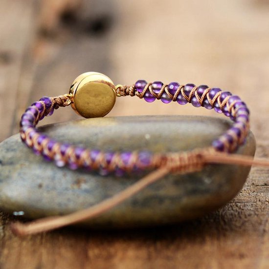 Gevlochten armband met paarse amethist kralen en de achterkant van een ronde Marly Quartz steen, liggend op een steen.