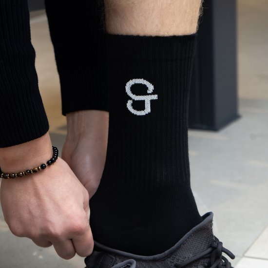 Zwarte sok met wit logo 'G'. Getoond op een persoon die de sok aantrekt, met sneakers aan.