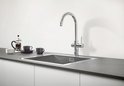 Chroom GROHE keukenkraan boven een metalen gootsteen, met bijpassende mokken en koffiezetter ernaast.