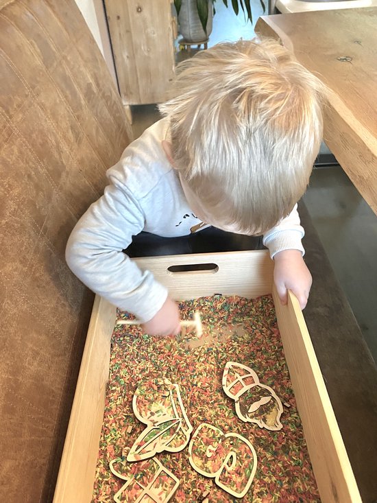 Jongetje met blond haar speelt met houten Sint Nicolaas thema figuren in een bak met gekleurde rijst.