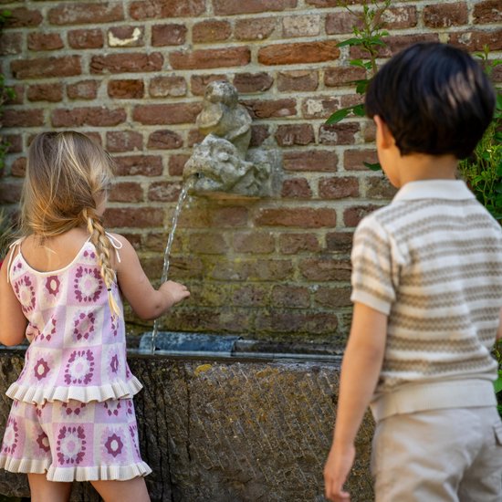 Meisje met blonde vlechtjes bij een fontein, draagt lila top met bloemenprint en ruches.