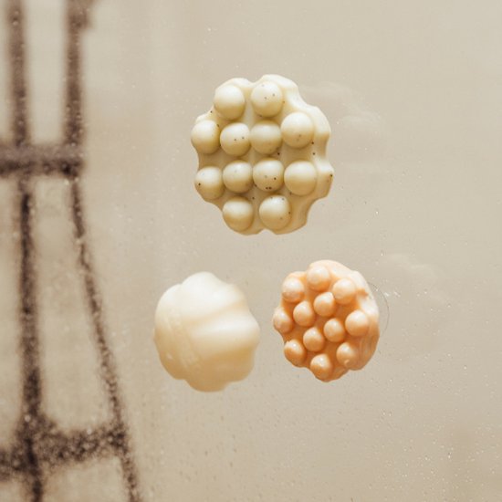 Drie Wondr shampoo bars op een natte douchewand: twee abrikooskleurige met nopjes, één romige variant.