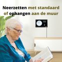 Oudere vrouw leest een boek, met op de muur een OER dementieklok die de datum 'woensdag 9 juni 2021' en tijd weergeeft. Tekst: 'Neerzetten met standaard of ophangen aan de muur'.