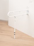 Opklapbare witte toiletbeugel met steunpoot, langs een witte muur.