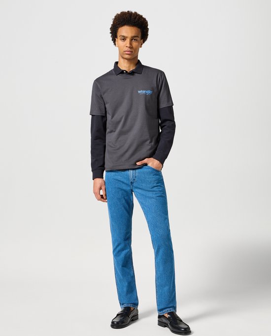 Model draagt een grijze Wrangler polo over een zwart shirt, met een blauwe jeans.