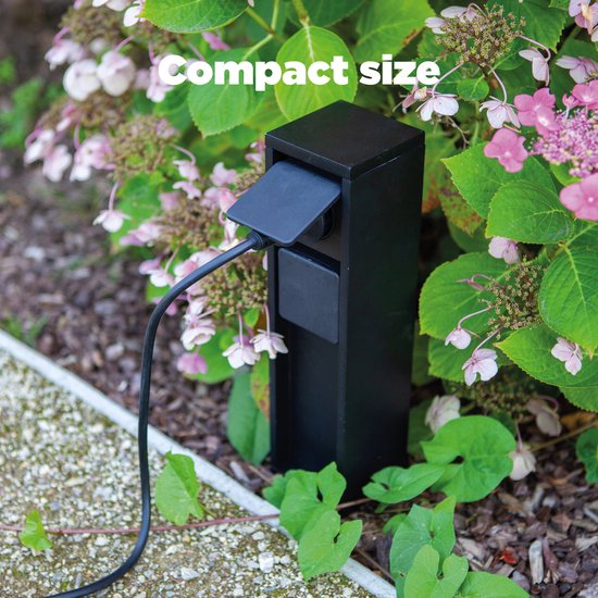 Zwarte buitenpaal met type E stopcontact in een compact formaat, omgeven door bloeiende planten.