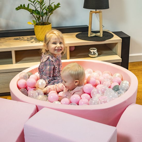 Twee kinderen lachen in een roze ballenbad met roze, witte en grijze ballen.