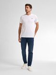 Man met de handen in de zakken, staand in een donkerblauwe, slimfit jeans en een witte T-shirt.