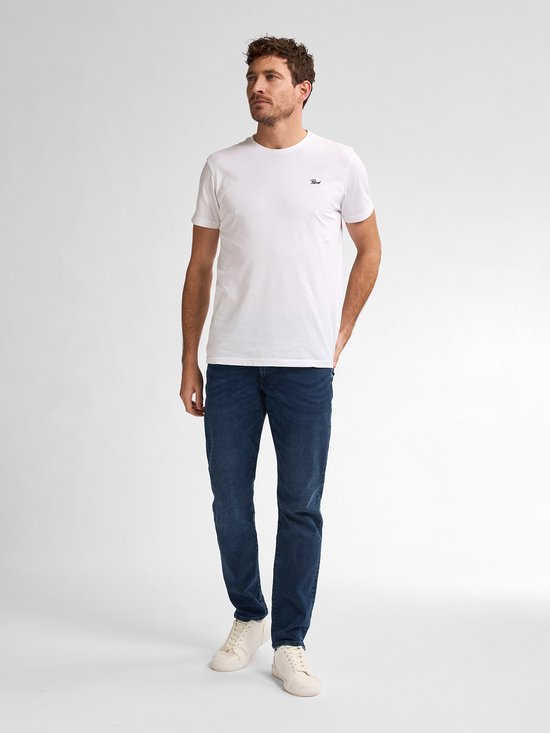 Man met de handen in de zakken, staand in een donkerblauwe, slimfit jeans en een witte T-shirt.