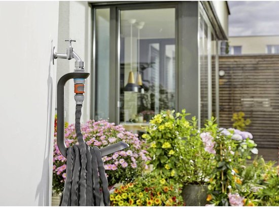Zwarte Gardena Liano TapFix slanghouder gemonteerd aan een witte muur met een tuinslang erin. Naast een kraan met aansluitstuk.