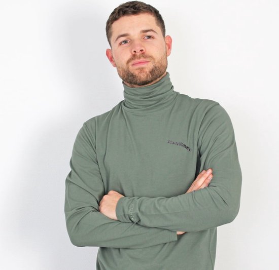 Man in een groene coltrui met lange mouwen, armen gekruist, tegen een witte achtergrond.