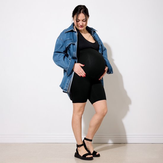 Zwangere vrouw in zwarte legging en denim jasje, met sandalen, staat bij witte muur.