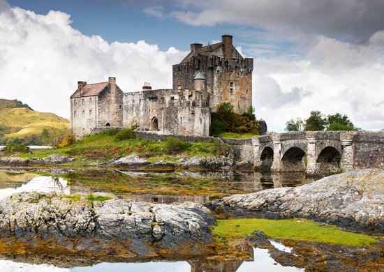 Kasteel Eilean Donan in Schotland met stenen brug en reflecties in het water, onder een bewolkte hemel.