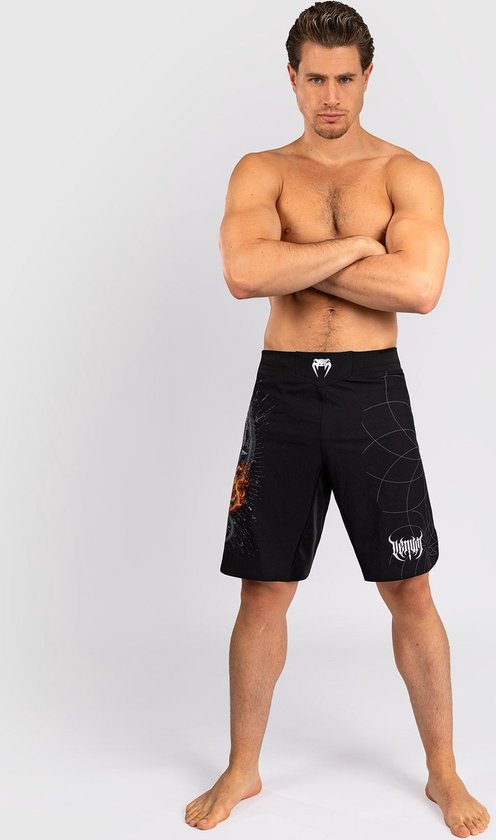 Man met gekruiste armen, draagt zwarte Venum sportbroek met vlammenprint op linkerbroekspijp en VENUM-logo op rechterbroekspijp.