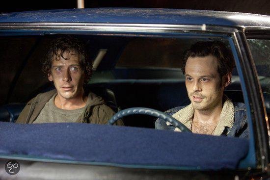 Twee mannen in een auto, één met een verwarde uitdrukking, de ander kijkt naar rechts.