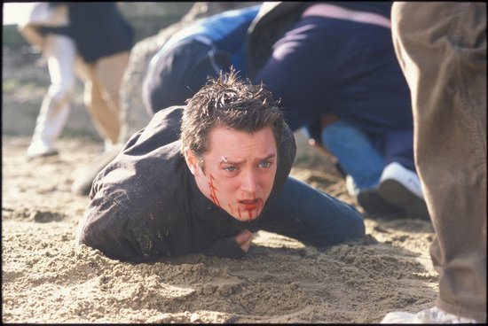 Elijah Wood met bloed op zijn gezicht, liggend op het zand, met blauwe ogen en een donkere jas.