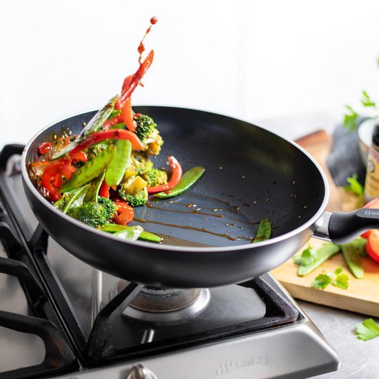 Zwarte wokpan op gasfornuis met spattende groenten en rode paprika. Handvat met "GREENPAN" logo.