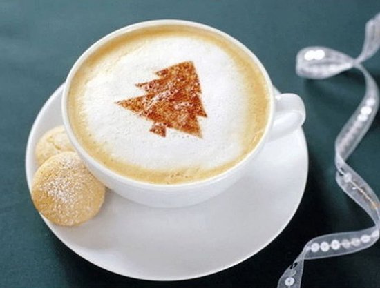 Koffiekopje met kerstboom-patroon op melkschuim, twee koekjes en een strik op de achtergrond.