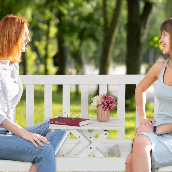 Twee vrouwen op een witte houten tuinbank met inklapbaar tafeltje, met een boek, bril en vaas met bloemen.