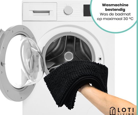 Een hand met een zwarte, zachte badmat bij een open wasmachine.