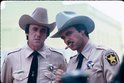 Twee mannen in beige sheriffuniformen met cowboyhoeden. De rechtse man rookt een sigaar.