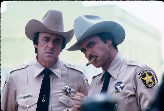 Twee mannen in beige sheriffuniformen met cowboyhoeden. De rechtse man rookt een sigaar.