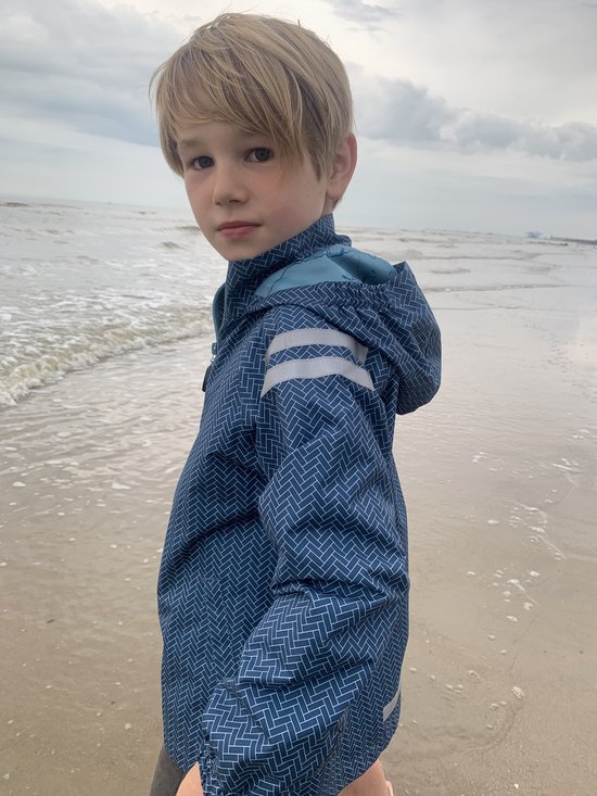 Jongen in een blauw regenpak met visgraatpatroon, staand op het strand.
