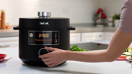 Een hand draait aan de knop van een zwarte Tefal multicooker met digitale weergave.