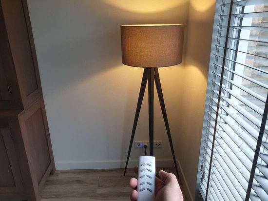 Een hand houdt een afstandsbediening vast bij een staande lamp met een stoffen kap en driepoot, bij een raam met jaloezieën.