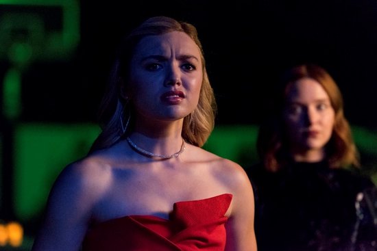 Een blonde vrouw met een rode off-shoulder jurk en een ketting, met een bezorgde uitdrukking.