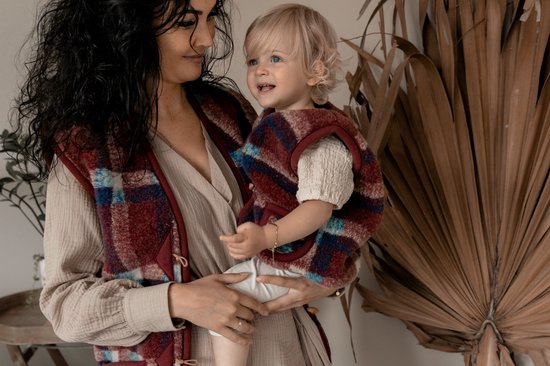 Vrouw houdt een baby vast, beiden dragen een bordeauxrode geruite bodywarmer met blauwe en witte ruiten.