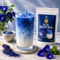 Glaasje Blue Matcha, ijsblokjes en melk met een blauwe poederverpakking ernaast. Blauwe vlinderbloemen op de voorgrond.