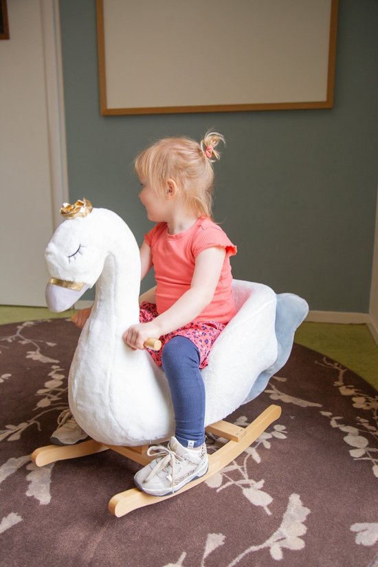 Een peuter in een oranje shirt en een roze rokje rijdt op een witte zwaan schommelstoel met gouden kroon.