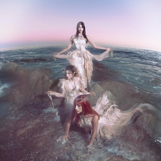 Drie vrouwen in lange, crèmekleurige jurken op rotsen in de zee, met een pastelkleurige lucht.