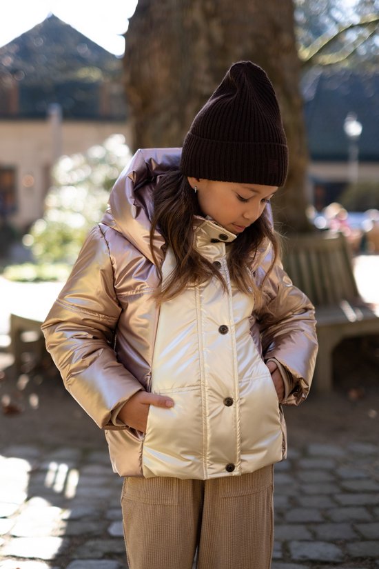 Meisje met bruine muts en roze/beige glanzende jas, handen in zakken.