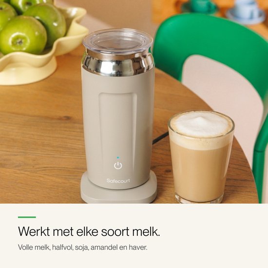 Beige melkopschuimer op tafel naast een glas met cappuccino. Een kom met groene appels is zichtbaar op de achtergrond.