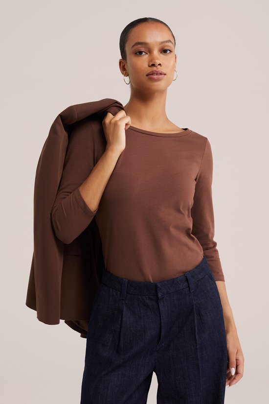 Model draagt een bruin T-shirt met driekwart mouwen en donkere broek, jasje over de schouder.