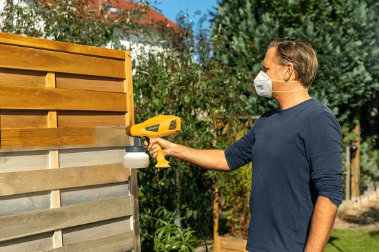 Man met mondkapje spuit houten hekwerk bruin met een geel Wagner verfspuitsysteem.