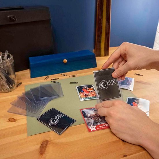 Handen van een persoon die trading cards in 'Eclipse' sleeves op een bureau legt, met andere kaarten en sleeves ernaast.