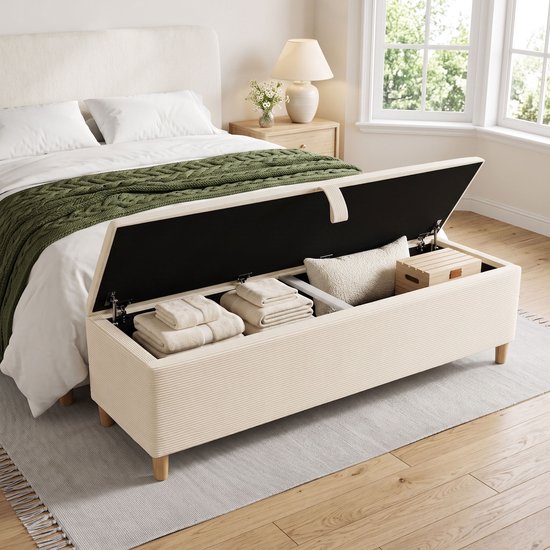 Banc de rangement beige en velours côtelé, ouvert révélant l'espace de rangement, à côté d'un lit dans une chambre.