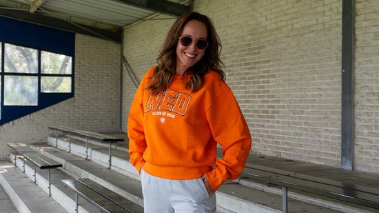 Vrouw met zonnebril draagt een oranje trui met "NED CLASS OF 2026" opdruk.