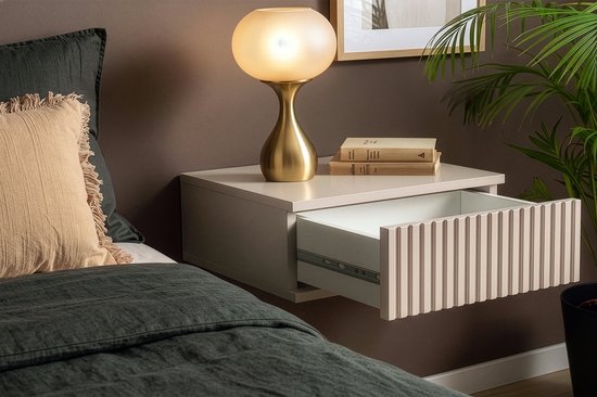Beige hangend nachtkastje met de lade open, verticale ribbels op het front, naast een bed met kussens.